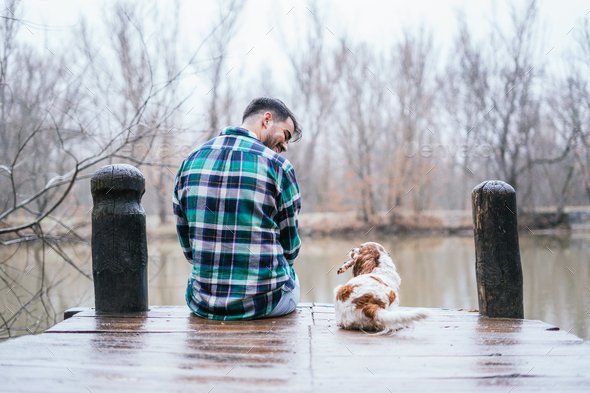 Smiling Hispanic man wearing flannel with Cavalier King Charles spaniel on a wooden dock in lake