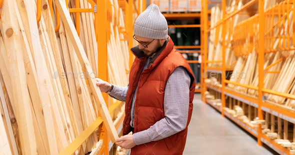 a male builder in a store chooses lumber among wooden bars and planks
