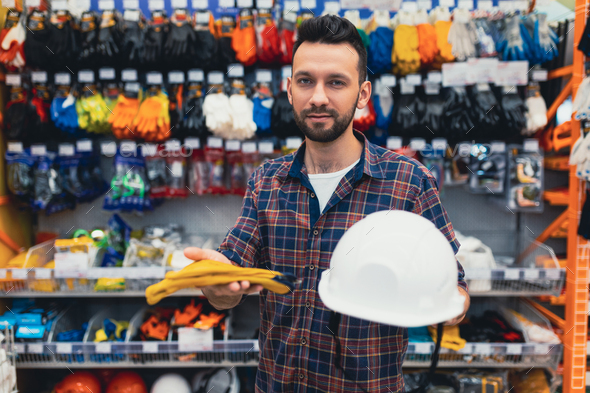 a male builder in a hardware store chooses a hard hat and protective gloves