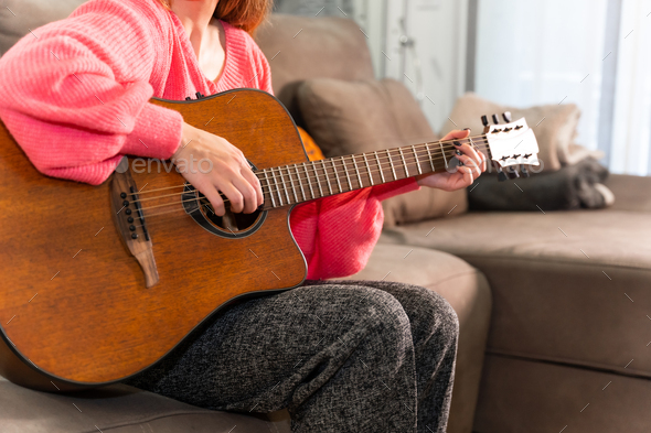 Woman playing the guitar at home sitting on the sofa in her living room next to the sale