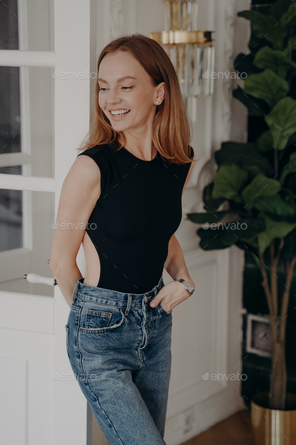 Attractive european lady in fashionable slim fit black bodysuit and jeans  looking aside and smiling. Stock Photo by StudioVK