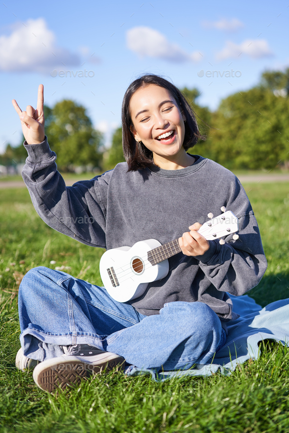 Happy girl playing ukulele in park, showing rock n roll, heavy metal sign and Stock Photo by