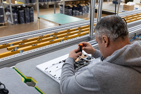 Photo of an adult man in a gray sweater who assembles a computer monitor system block on an conveyor