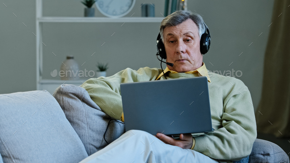 Senior 60s male on couch at home wear digital earphones talking online to laptop web camera old man