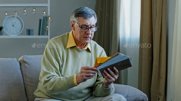 Old 60s elderly Caucasian man checking sorting letters in living room concentrated mature