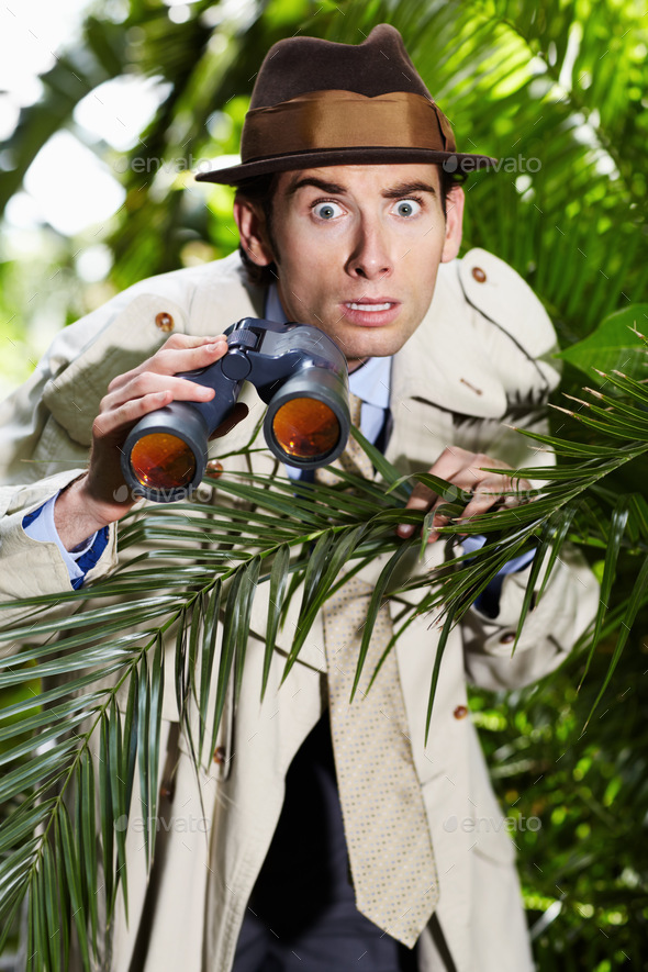 Shocked private investigator using binoculars to spy on someone from the bushes