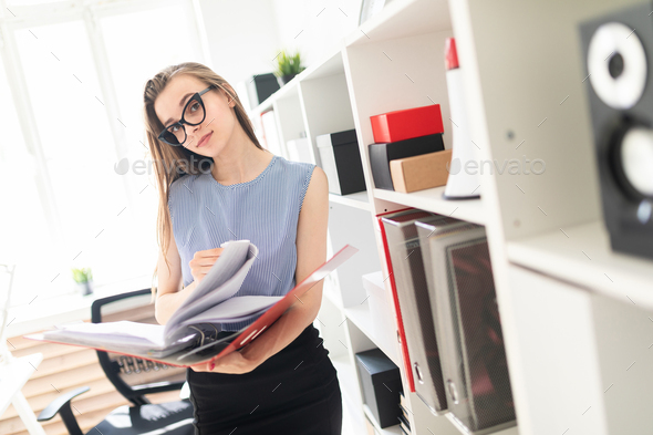 Beautiful young girl in an office stands near a rack and scrolls through a folder with documents.