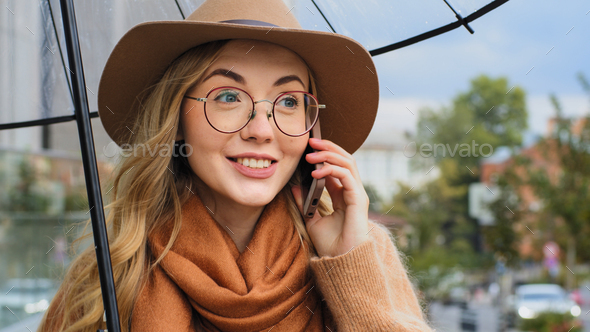 Attractive young woman with umbrella answers call on the street during rain, smart girl communicates