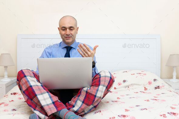 Man working with a laptop in a bed wearing a shirt and tie and pajama pants
