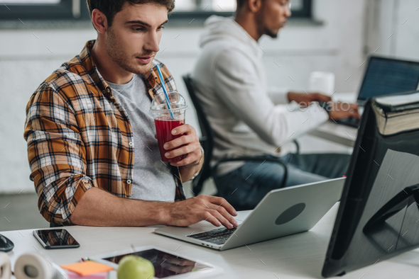 selective focus of young programmer drinking juice while working near ...