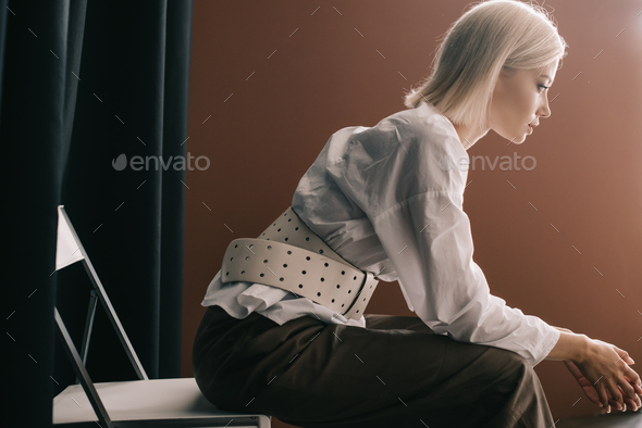 side view of stylish blonde woman in white blouse sitting on chair near curtain on brown