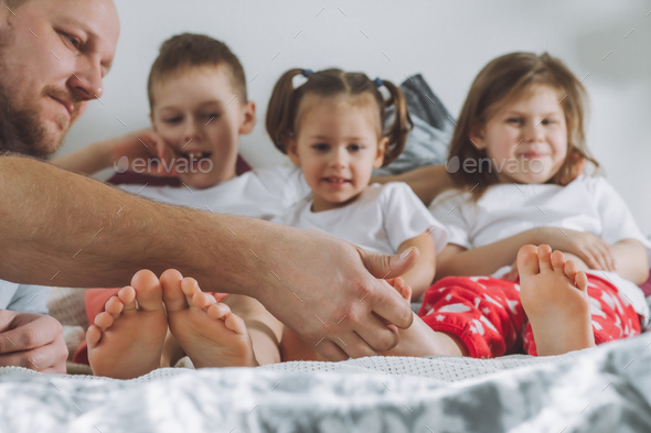 Father plays with three children on bed. Dad tickles kids feet. Family of daddy, two girls, and boy
