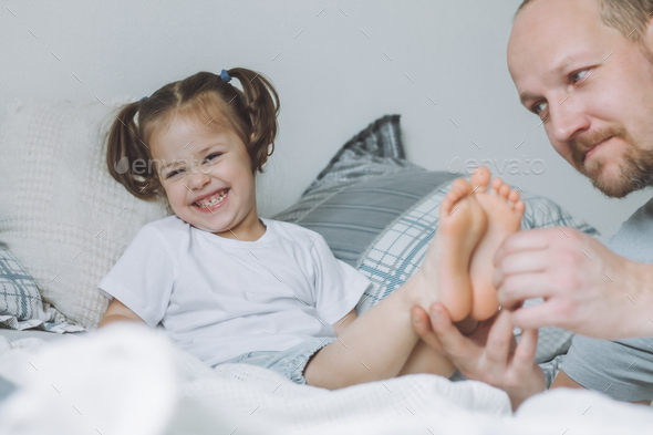 Father plays with little daughter 2-4 on bed. Dad tickles kids feet. Family, having fun