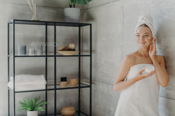 Photo of relaxed young woman applies face cream, has satisfied expression