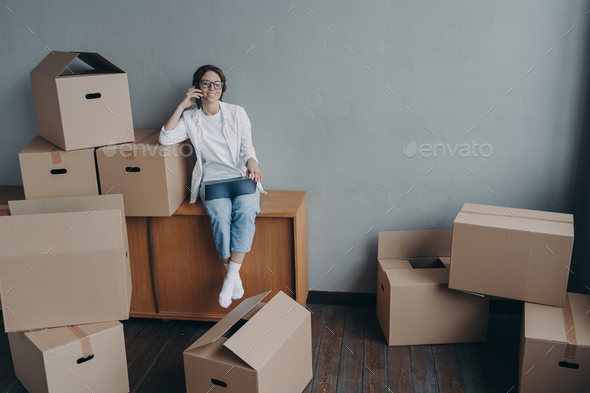 Female calling moving company, orders mover for relocation, sitting with laptop and cardboard boxes