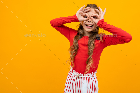 surprised girl with curly hair in a red blouse and striped trousers peers through the palms on an