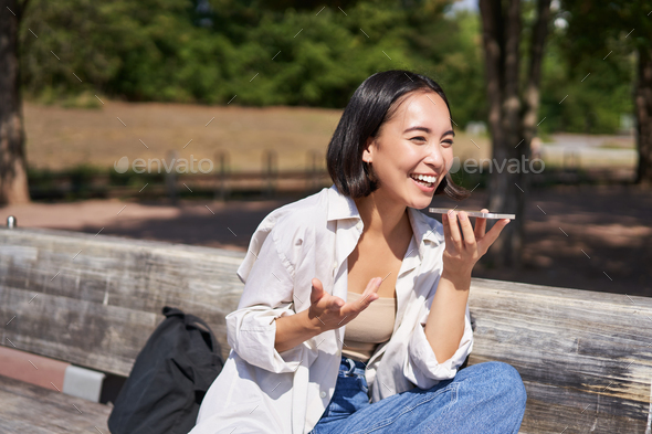Portrait of happy asian girl records voice message, talking via speakerphone on smartphone, sitting