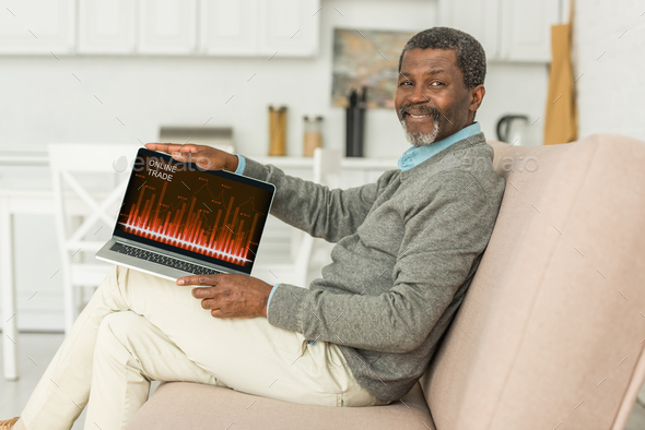 smiling african american man sitting on sofa and showing laptop with online trade app on screen