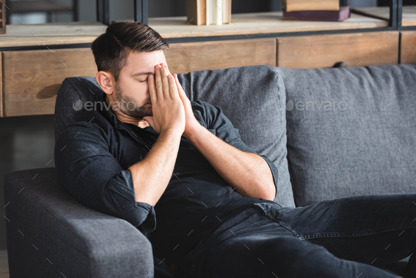 man with headache sitting on sofa and obscuring face in apartment