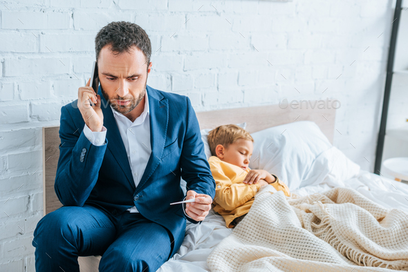 worried man in formal wear talking on smartphone and holding thermometer while sitting on bed near