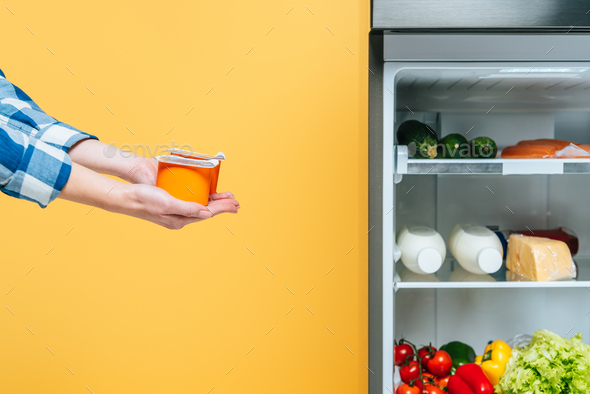 cropped view of woman holding yogurt near open fridge with fresh food on shelves isolated on yellow