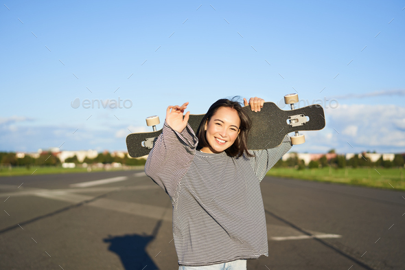 Leisure and people. Happy asian woman standing with longboard, cruising on an empty road in