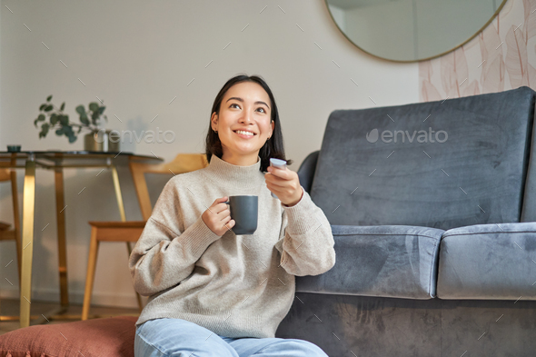 Portrait of smiling korean woman sitting near tv, holding remote and ...