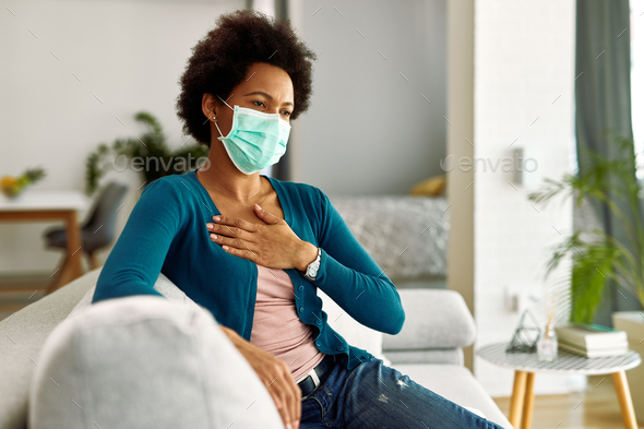 African American woman with face mask feeling chest pain while sitting at home.