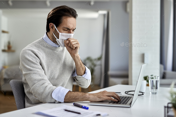 Businessman with protective face mask coughing while working on laptop at home.