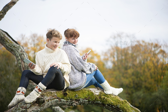 mother and son teen sit with their backs together on a tree branch and use cell phones and headsets