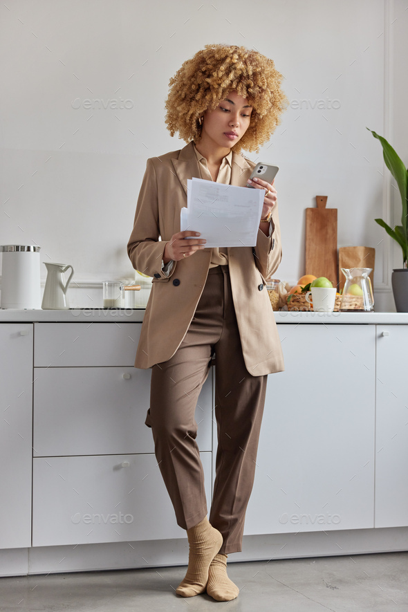 Serious woman holds papers and smartphone studies paper bills tries to deal with payments online