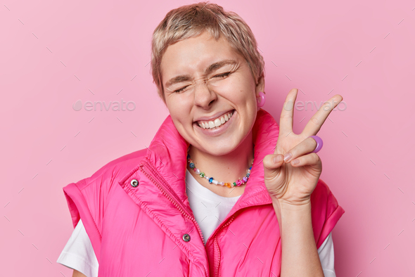 Positive young European woman with short hairstyle makes peace gesture smiles toothily keeps eyes