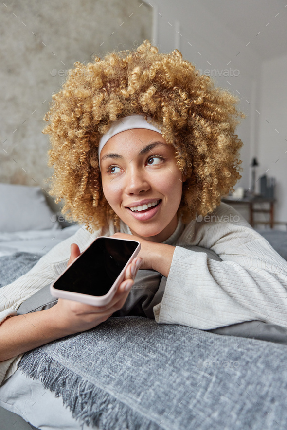Happy relaxed curly haired woman lies on comfortable bed holds mobile phone near mouth records voice