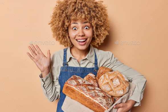 Surprised curly haired female baker works in bakehouse keeps hand raised smiles broadly poses with