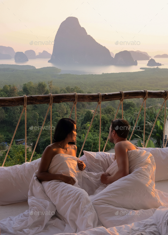 couple waking up in bed looking out over ocean during sunrise at wooden hut moutains of Thailand
