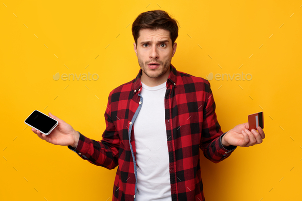 Confused man holding credit card and smartphone, shrugging