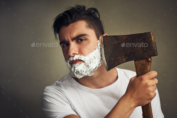 Its the right tool for the job. Shot of a handsome young man shaving his beard with a lumberjack.