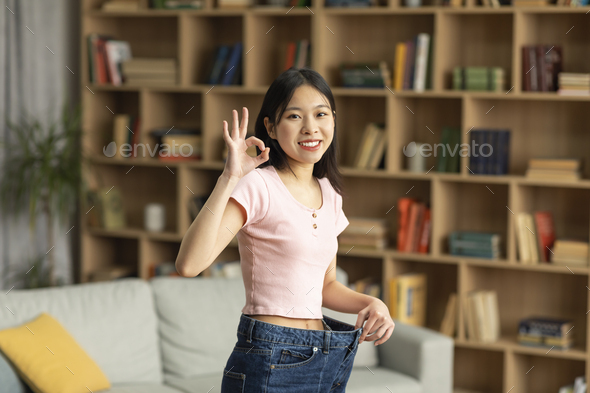 Happy asian lady showing result of successful weight loss, gesturing ok and wearing old oversize