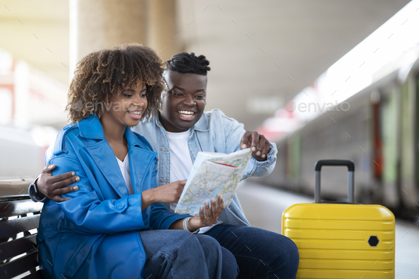Black Couple Checking Travel Route On Map While Sitting At Railway Station