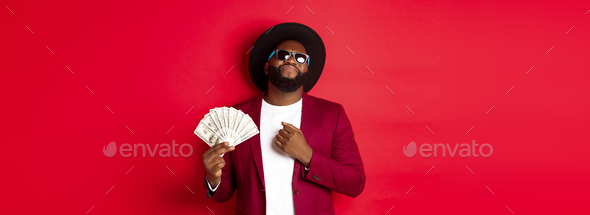 Sassy and cool african american man in sunglasses and hat, pointing at himself and showing dollars