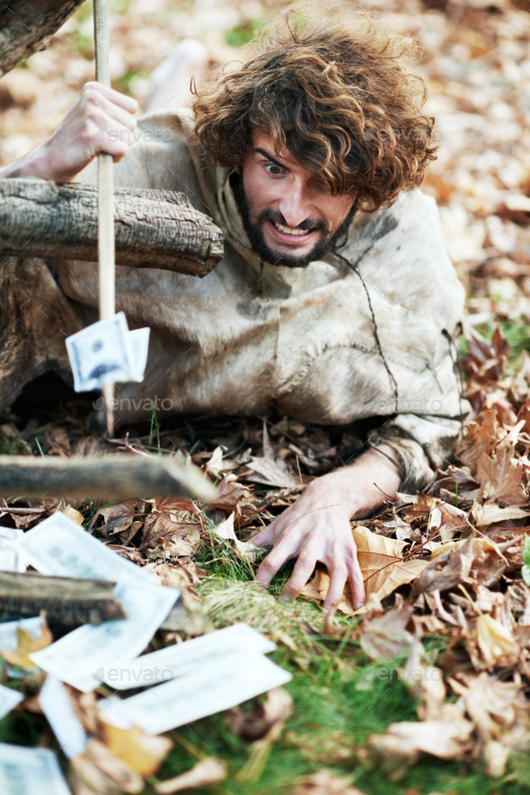 A crazy looking caveman crawling on the ground and piercing money with his spear