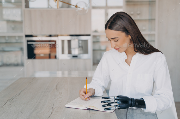 Modern disabled girl with bionic prosthesis hand writing in notebook, sitting at table at home