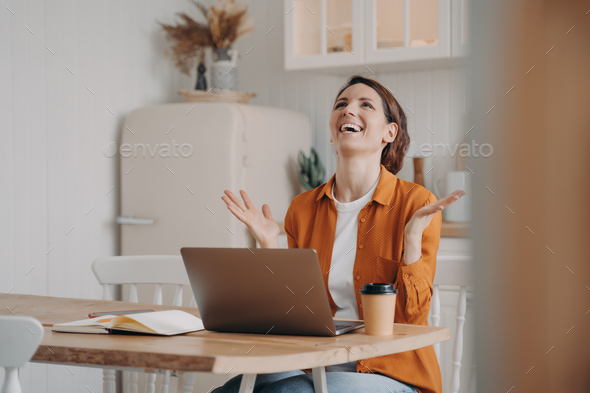 Happy hispanic woman got email with perfect good news, gesturing, sitting at laptop in kitchen