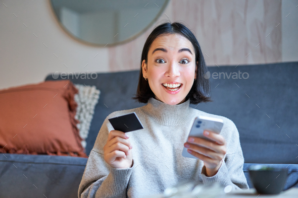 Enthusiastic girl sits at home with smartphone and credit card, pays for online order, book tickets