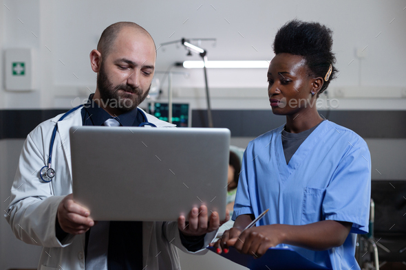 Doctor holding laptop presents patient medical history to nurse holding clipboard with lab results