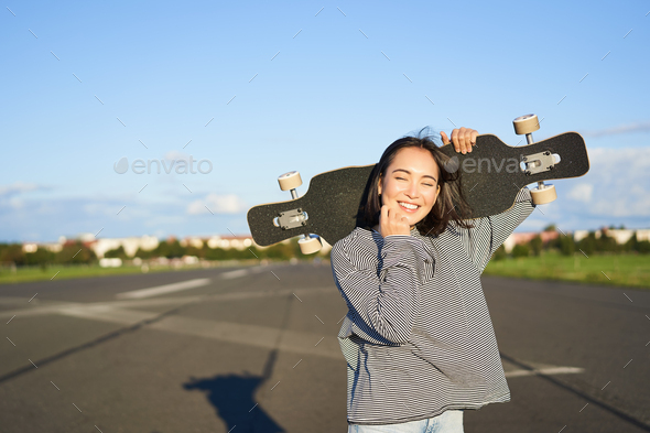 Leisure and people. Happy asian woman standing with longboard, cruising on an empty road in