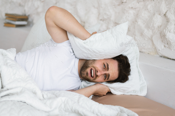 Tired angry sad young caucasian guy lies on bed, covers his ears with pillow, suffers from noisy