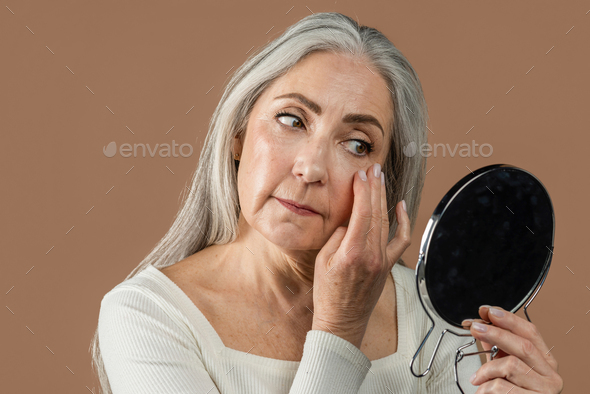 Serious elderly caucasian lady with gray hair looks in mirror, touches wrinkles, applies cream