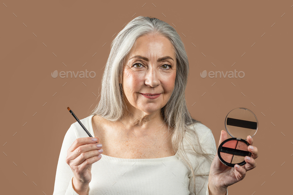 Cheerful senior european lady with gray hair shows cosmetics, blush or eye shadow with brush