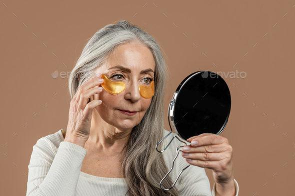 Serious elderly caucasian woman with natural beauty applies patches under her eyes and looks in